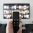 A person holds a remote control in front of a TV displaying a streaming platform interface with various movie and show thumbnails. The room includes a desk with two small lamps on either side of the TV.