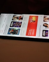 A smartphone displaying a streaming app interface, showing various movie and TV show thumbnails. The screen includes categories like 'For you' and 'New release hit films', with images from popular films. The background is a dimly lit setting with a fabric surface.