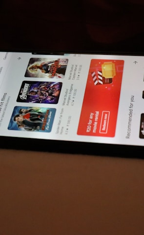 A smartphone displaying a streaming app interface, showing various movie and TV show thumbnails. The screen includes categories like 'For you' and 'New release hit films', with images from popular films. The background is a dimly lit setting with a fabric surface.