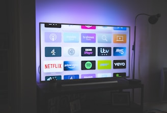 A flat-screen television is displayed in a dimly lit room, showcasing a variety of streaming service apps on its screen, such as Netflix, Prime Video, and Spotify. The TV is positioned on a black stand beside a tall floor lamp, casting a soft ambient light on the surrounding area.