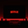 A television screen displays the Netflix logo in red against a black background. The room is dimly lit with a red glow possibly from LED backlighting behind the screen. Two speakers are positioned on either side of the TV set atop a minimalist entertainment center.