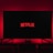 A dark room featuring a television screen displaying the word 'Netflix' in red against a black background. The screen is centered on a console table, with a video game controller placed in front. The room is dimly lit by red ambient lighting, creating a cozy atmosphere.