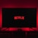 A dark room featuring a television screen displaying the word 'Netflix' in red against a black background. The screen is centered on a console table, with a video game controller placed in front. The room is dimly lit by red ambient lighting, creating a cozy atmosphere.