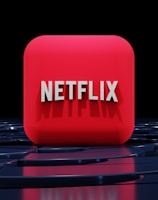 A red, cube-shaped icon with the word 'NETFLIX' prominently displayed in white, bold letters. The cube is set against a dark, glossy, circular patterned background, reflecting a sleek, futuristic design.