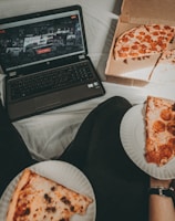 A cozy setting with a laptop displaying a streaming service interface nearby, alongside a pizza box holding a partially eaten pizza. Two slices of pizza are on paper plates held by someone, suggesting a casual movie night atmosphere.