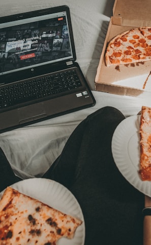 A cozy setting with a laptop displaying a streaming service interface nearby, alongside a pizza box holding a partially eaten pizza. Two slices of pizza are on paper plates held by someone, suggesting a casual movie night atmosphere.