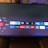 A television screen displays a smart TV interface with multiple app icons arranged in rows. Options such as Settings, Sources, Home, Search, and TV are listed on the left side. Some recognizable app icons include Netflix, Prime Video, BBC iPlayer, ITV Hub, All 4, My5, YouTube, and TikTok. The time and date are shown in the top right corner.