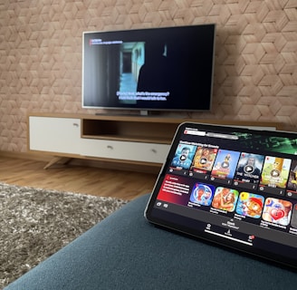 A modern living room features a flat-screen television mounted on a light-colored wooden stand with a patterned wall backdrop. Below the TV, a carpet lies on a wooden floor. In the foreground, a tablet displays a streaming platform interface with various show thumbnails, suggesting a relaxed entertainment setup.